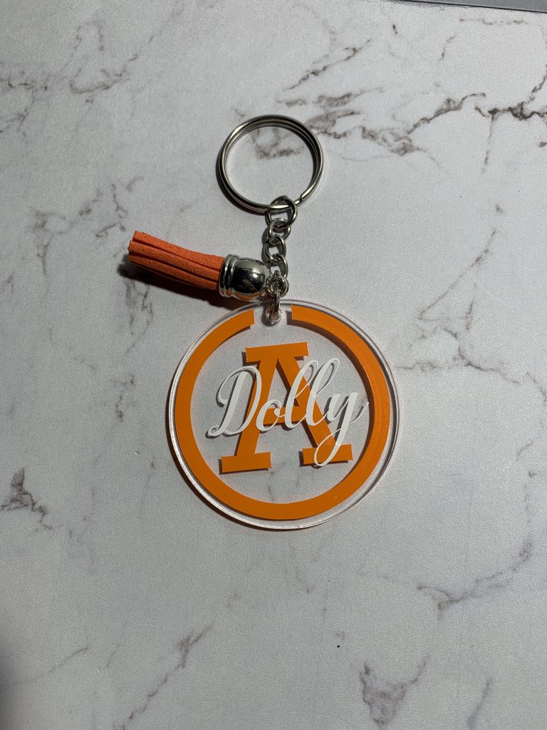 Circular keyring with name. Featuring orange outline, initial and tassel and finished with white name.