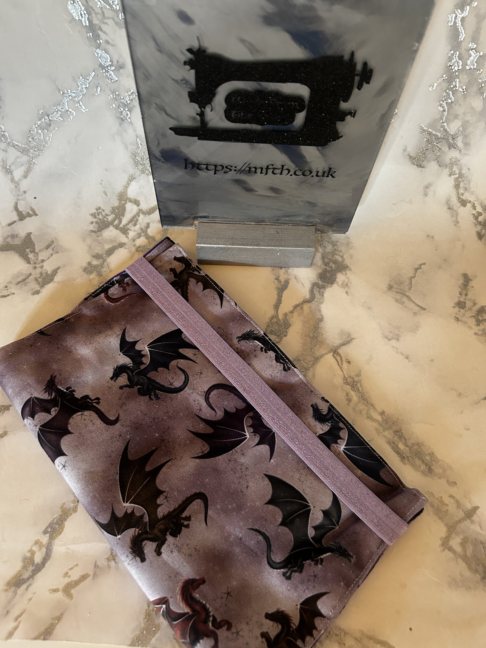 Book Cover handmade from fabrics with an elasticated band to hold it together around the top. The print is purple and grey mix on the background with colourful dragons flying around on it