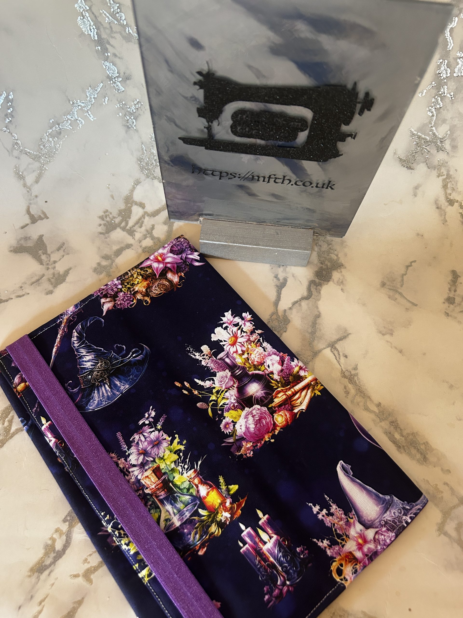 Book Cover in fabric with an elasticated band around the edge. The print is dark purple black with witches hats flowers, potion bottles and more on it