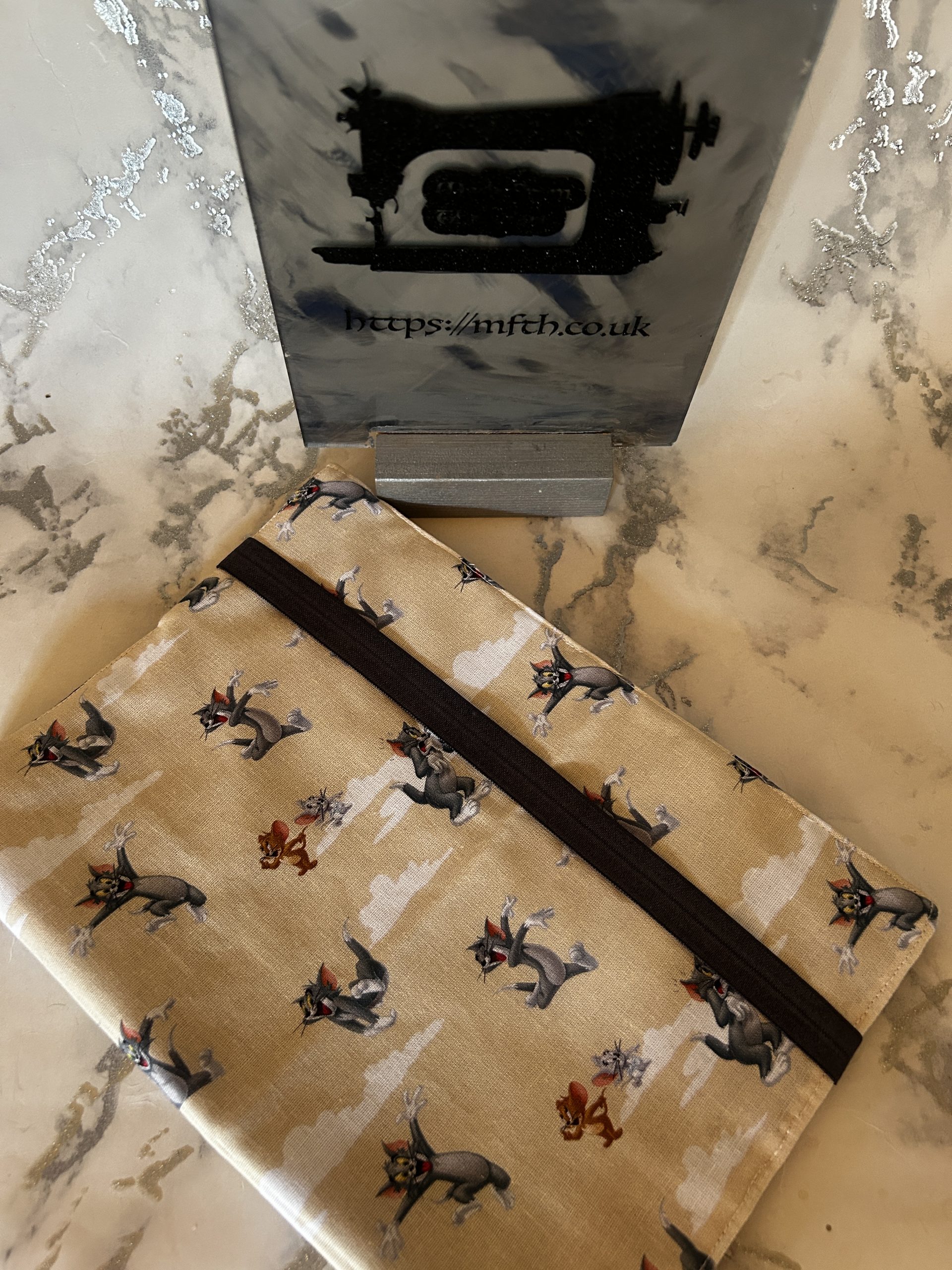 Book cover in fabric with an elasticated edge on the side. The print is beige featuring everyones favourite Tom and Jerry